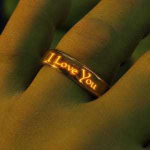 Könnte beinhalten: Ein metallischer Ring mit einer glatten, polierten Oberfläche und dem eingravierten Text I Love You entlang des Bandes. Die gravierten Buchstaben strahlen ein warmes, leuchtendes Orange aus, das einen Kontrast zum dunklen Metall bildet.