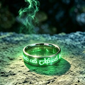 May include: A metallic ring featuring glowing green inscriptions along its outer band, resting on a textured, dark stone surface with wisps of green smoke rising from the surrounding area, creating a luminous effect on the rock.