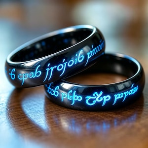 May include: Two polished black metal rings featuring glowing blue engraved script lettering that wraps around the outer surface of each band. The rings have a smooth, reflective finish and are positioned overlapping each other in a close-up view.