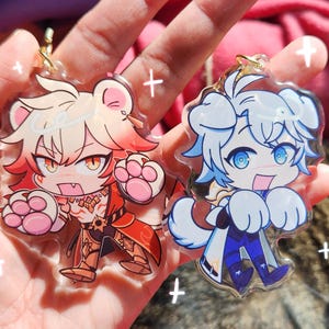 May include: Two clear acrylic keychains featuring anime-style characters. The left keychain depicts a character with blonde hair, bear ears, and red clothing. The right keychain shows a character with white hair, dog ears, and blue clothing.