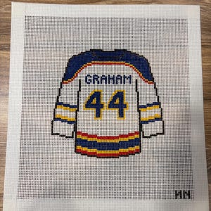 May include: A needlepoint canvas featuring a white hockey jersey design with blue shoulders and sleeves. The jersey displays the name GRAHAM in blue letters above the number 44 in blue with yellow outlines. Horizontal blue, yellow, and red stripes accent the sleeves and hem.