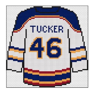 Könnte beinhalten: Kreuzstich-Design eines Hockey-Trikots. Das weiße Trikot hat blaue und goldene Akzente. Der Name "TUCKER" steht über der Nummer "46" in Blau. Das Design ist ein pixeliges, handgefertigtes Kunsthandwerk.