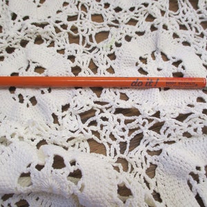 May include: An orange pencil with the text "do it!" and "REDDY KILOWATT" printed on the side. The pencil is lying on a white crocheted lace doily with a floral pattern.