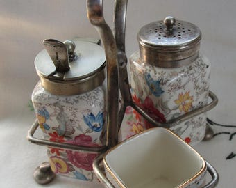 May include: A vintage condiment set featuring a metal stand holding two floral-patterned jars with silver lids and a small square dish. The jars and dish have a white background with colorful floral designs. The metal stand has a curved handle.