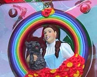 May include: A round Christmas ornament featuring Dorothy from The Wizard of Oz, holding Toto, framed by a rainbow. The ornament has a red and yellow floral base and a gold string for hanging. The text "Dorothy & Toto" is on a green tag.
