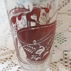 May include: Clear glass tumbler with a maroon design. The design features a stylized boat, floral elements, and a bird. The glass has a slightly flared shape and is likely vintage or antique.