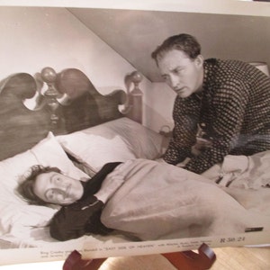 May include: A black and white vintage film still featuring two individuals in a bedroom setting. One individual lies in a bed with a dark top, while another stands over the bed wearing a patterned long-sleeved shirt. Text at the bottom reads: R 30 24.