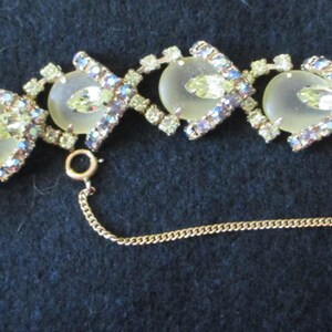 May include: A vintage bracelet featuring a gold-tone metal chain with alternating oval and diamond-shaped links. The oval links have a pale yellow center stone, surrounded by clear and iridescent rhinestones. The bracelet has a safety chain and a clasp.