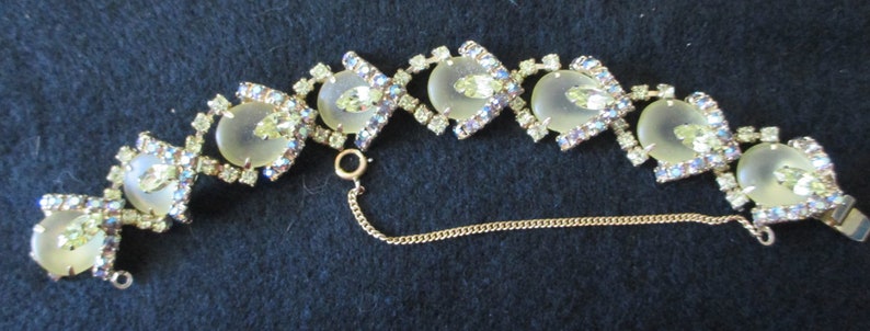 May include: A vintage bracelet featuring a gold-tone metal chain with alternating oval and diamond-shaped links. The oval links have a pale yellow center stone, surrounded by clear and iridescent rhinestones. The bracelet has a safety chain and a clasp.