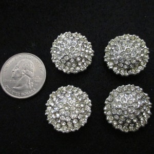 May include: Four silver-toned round buttons with a raised design of clear rhinestones. The buttons are approximately the size of a US quarter.