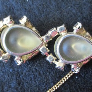 May include: Close-up of a gold-toned necklace featuring pear-shaped, light-colored stones. Each stone is surrounded by small, clear crystals. The necklace is on a dark background.