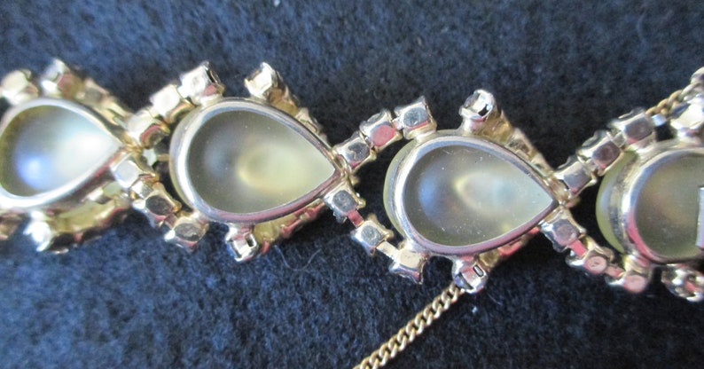 May include: Close-up of a gold-toned necklace featuring pear-shaped, light-colored stones. Each stone is surrounded by small, clear crystals. The necklace is on a dark background.