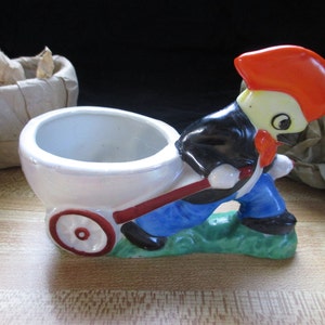 May include: A ceramic figurine of a cartoon character pushing a wheelbarrow. The character is wearing a black jacket, blue pants, and a red hat. The wheelbarrow is white with a red handle and a red wheel.