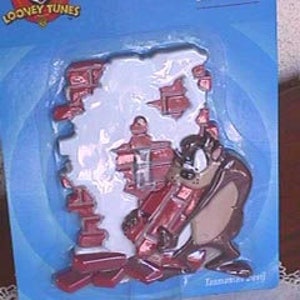 May include: A Looney Tunes Tazmanian Devil switch plate in a blue package. The plate is shaped like a hole in a wall with Taz emerging from the side. The package has the Looney Tunes logo and the GE logo.