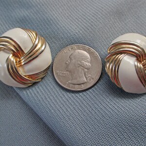 May include: A pair of vintage clip-on earrings. Each earring features a white, round, enameled center encircled by gold-tone metal bands. A U.S. quarter coin is placed between the earrings for size comparison. The earrings are set against a light blue fabric background.