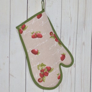 May include: A beige oven glove with a green trim and a hanging loop. The glove features a pattern of red strawberries and green leaves, with cursive script. It hangs against a white wooden backdrop.