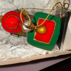 May include: Two Christmas ornaments in a box. One is a red circle, the other is a green square with a red center. Both have gold accents and gold hanging cords. The ornaments are in a box with white paper.
