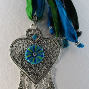 May include: A silver heart-shaped pendant with a blue and green evil eye charm hangs from a turquoise and green silk ribbon necklace.