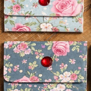 May include: Two rectangular fabric wallets featuring floral patterns. The top wallet is light blue with pink roses and white flowers. The bottom wallet is dark blue with pink roses and white flowers. Each wallet has a single red circular button.
