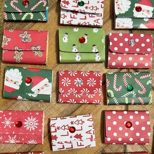 May include: A collection of small rectangular paper envelopes featuring various Christmas patterns including Santa Claus, candy canes, gingerbread men, snowmen, snowflakes, and the text hope peace and fa la la, each adorned with a small colored gem.