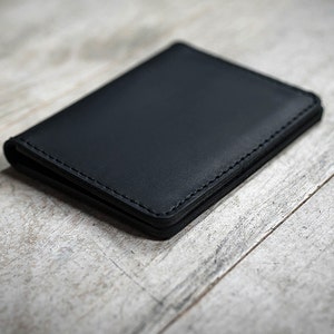 May include: A black leather wallet with visible stitching. The wallet is rectangular and appears to be closed. The background is a light-colored, textured wooden surface.