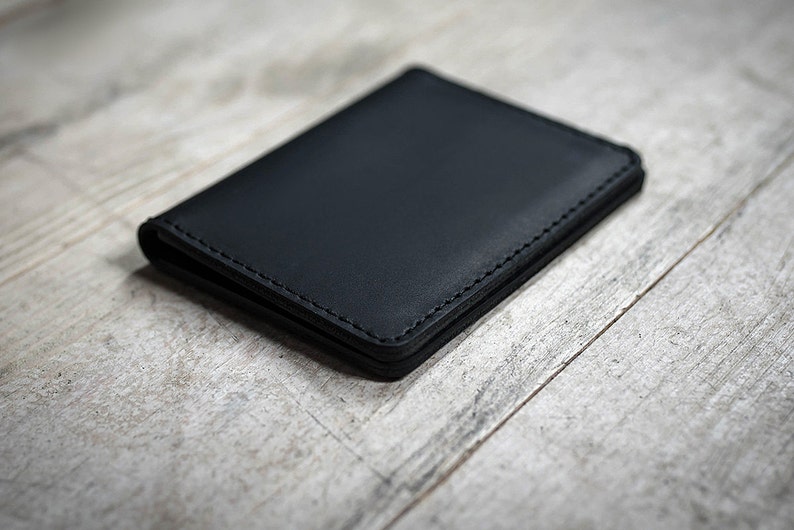 May include: A black leather wallet with visible stitching. The wallet is rectangular and appears to be closed. The background is a light-colored, textured wooden surface.