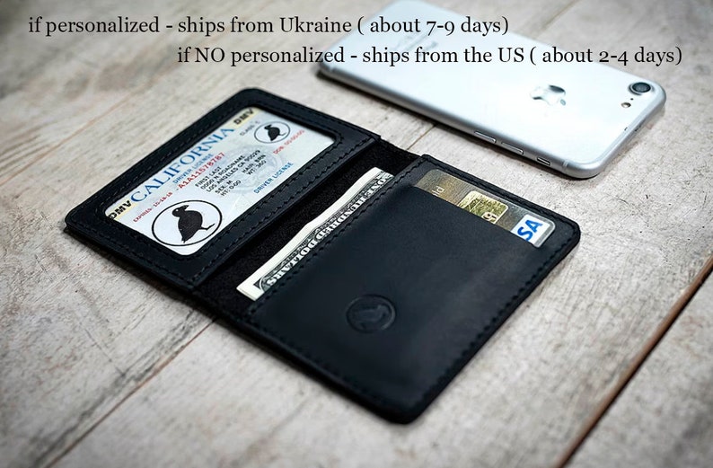 May include: Black leather wallet with a California driver's license visible. The wallet is open, displaying a dollar bill and a credit card. A smartphone rests in the background. The wallet has a small emblem on the front.