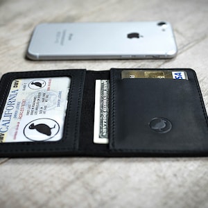 May include: Black leather wallet with a driver's license, cash, and credit cards visible. The wallet is open, displaying its contents. A silver smartphone rests in the background. The wallet has a debossed logo.