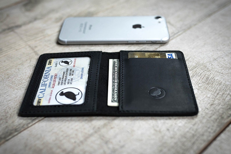 May include: Black leather wallet with a driver's license, cash, and credit cards visible. The wallet is open, displaying its contents. A silver smartphone rests in the background. The wallet has a debossed logo.