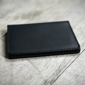 May include: A black leather wallet with stitched edges, resting on a light-colored wooden surface. The wallet is rectangular and appears to be empty. The stitching is a lighter shade of black.
