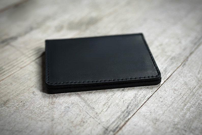 May include: A black leather wallet with stitched edges, resting on a light-colored wooden surface. The wallet is rectangular and appears to be empty. The stitching is a lighter shade of black.