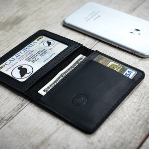 May include: Black leather wallet with a California driver's license and a dollar bill visible. A credit card is also in the wallet. A silver smartphone rests on the wooden surface next to the wallet.