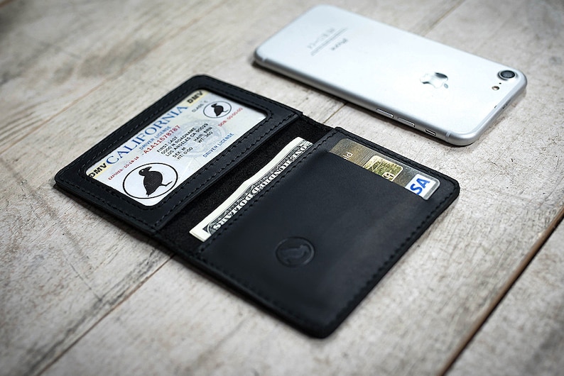 May include: Black leather wallet with a California driver's license and a dollar bill visible. A credit card is also in the wallet. A silver smartphone rests on the wooden surface next to the wallet.