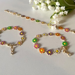 May include: Three gold chain bracelets with colorful daisy flower charms. The bracelets are on a white surface.