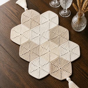May include: A handmade, cream and tan crochet table runner with a geometric design. The runner features a series of connected hexagonal shapes and is adorned with tassels at each end. Two crystal wine glasses and a small vase with wheat stalks are also on the table.