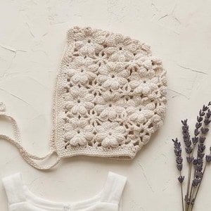 Könnte beinhalten: Draufsicht auf Babykleidung und Accessoires. Ein weißer, gestreifter Strampler, eine gehäkelte Mütze mit floralen Details, eine Holzrassel und ein Bündel Lavendelzweige sind auf einer cremefarbenen Oberfläche angeordnet. Drei kleine, glatte Steine sind ebenfalls vorhanden.