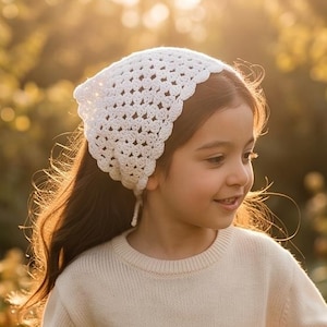 May include: A young person wearing a cream-colored sweater, matching pants, and a white crocheted head covering. They are holding a small woven basket. The background is a sunlit garden with blurred greenery.
