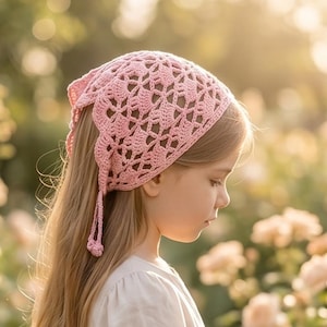 May include: A young person wearing a light pink, open-weave crochet headscarf with long, flowing hair. The headscarf has a decorative edge and ties. The person is wearing a light-colored dress, standing in a garden with soft, blurred flowers.