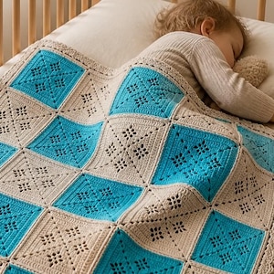 Peut inclure: Une couverture au crochet faite à la main sur un lit bébé. La couverture présente un motif carré avec des carrés turquoise et beiges alternés. Elle a un bord festonné décoratif. Un enfant dort sous la couverture.