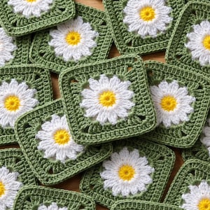 Puede incluir: Cuadrados de ganchillo hechos a mano con diseño de margarita. Cada cuadrado presenta una margarita blanca con centro amarillo sobre un fondo verde. Los cuadrados están colocados sobre una superficie de madera.