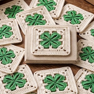 May include: A collection of cream-colored, square crochet coasters with green four-leaf clover designs. The coasters are arranged on a wooden surface, with a small stack of coasters and a ball of green yarn visible.