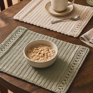 May include: Two rectangular crocheted placemats, one in cream and one in sage green, featuring a ribbed center texture and decorative border patterns. The cream placemat holds a ceramic mug on a saucer with a spoon, and the green one holds a bowl.