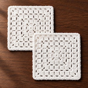 May include: Two square coasters made of off-white yarn, each featuring a detailed crochet granny square pattern with a repeating grid of small, open-work stitches and a solid, uniform border edge that frames the entire square shape.