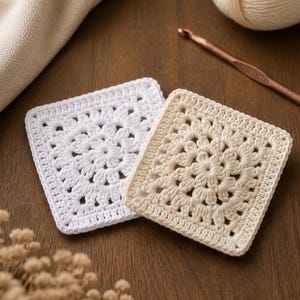 Puede incluir: Dos posavasos cuadrados de ganchillo en blanco y crema. Los posavasos presentan un diseño floral en el centro y un borde sólido. Un ovillo de lana crema y un ganchillo se ven en el fondo.