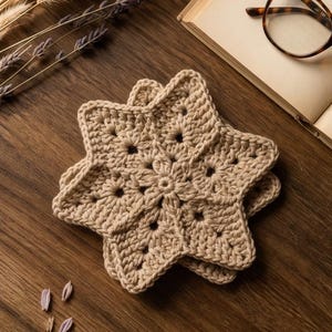 Könnte beinhalten: Ein Stapel aus zwei beigen, sternförmigen Häkeluntersetzern mit einem zentralen Blumenmuster und durchbrochenen Details. Die Untersetzer sind aus Garn gefertigt und weisen eine strukturierte Maschenkonstruktion mit sechs markanten Spitzen auf.