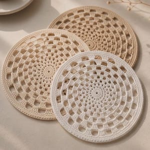 Puede incluir: Tres posavasos circulares de ganchillo en varios tonos de blanco y beige. Cada posavasos presenta un patrón geométrico detallado y repetitivo con un punto central y puntadas caladas radiantes que crean una textura de encaje.