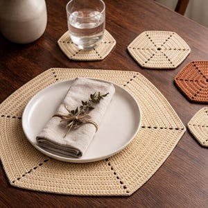 Può includere: Tavola apparecchiata con un runner e sottobicchieri esagonali beige all'uncinetto. Un piatto bianco con un tovagliolo di lino e rametti verdi è posto sul runner. Sono presenti anche un bicchiere d'acqua e un vaso con fiori secchi.