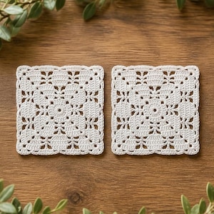 May include: Two square, off-white crochet coasters with a floral design. The coasters are placed on a wooden surface, with green foliage in the background. The coasters are approximately 4 inches square.