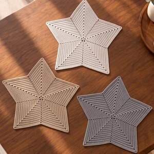 May include: Three star-shaped crochet mats in light beige, tan, and grey. Each mat features a dense, textured stitch pattern radiating from the centre to the five points, creating a uniform geometric design with a clean, finished edge.