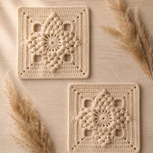 Puede incluir: Dos posavasos cuadrados de ganchillo color crema. Cada posavasos presenta un diseño central en forma de estrella con detalles texturizados en relieve y cortes cuadrados. Los posavasos están colocados sobre una superficie de madera clara, con hierba ornamental seca.